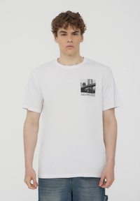 T-shirt di cotone bianco con maniche corte, caratterizzato da una stampa fotografica in bianco e nero sulla parte sinistra del petto e un collo rotondo.