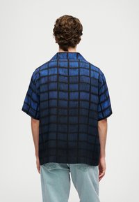 Mann med krøllete brunt hår som har på seg en blå og svart rutet kortermet skjorte og lyseblå jeans, står med ryggen til kameraet.