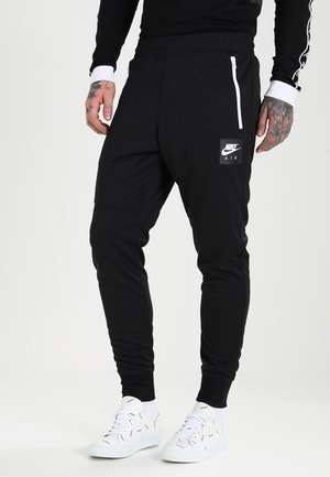 Man die zwarte Nike Air joggers draagt met een witte ritssluiting op de zak en witte sneakers met zwarte kronkelpatronen, staand op een witte achtergrond.