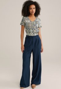 Ziedu raksta, īsām piedurknēm, V veida kakla augšdaļa krēmkrāsā ar zilām akcentiem, apvienota ar platām kājām, tumši zilo biksēm un miesa krāsas augstpapēžu kurpēm.