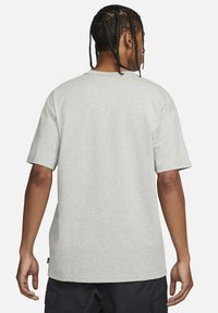Persona ar pītām matēm, tērpusies vienkāršā gaiši pel�ēkā īsā piedurkņu t-shirt un melnās kargo biksēs, stāv pretēji balta fona.
