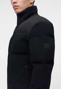 Homme portant une doudoune noire côtelée avec un col montant et un patch à l’épaule montrant le logo "BOSS" sur le haut du bras gauche.