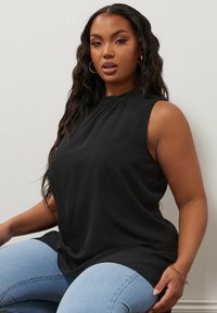 Top nero senza maniche con collo alto, tessuto morbido, design drappeggiato e pieghe sottili sul colletto. Abbinato a jeans azzurri chiari.
