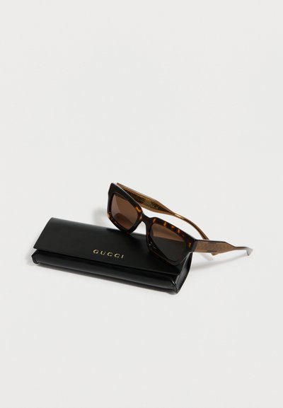 Τετράγωνα γυαλιά ηλίου με καφέ φακούς και σχέδιο χελώνας, τοποθετημένα πάνω σε μαύρη θήκη Gucci με χρυσά γράμματα, σε λευκό φόντο.