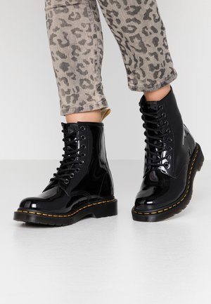 Osoba nosząca błyszczące czarne, sznurowane buty wojskowe i szare, krótkie spodnie w panterkę stojąca na białej podłodze.