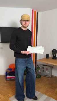 Model trägt ein schwarzes Langarmshirt und weit geschnittene blaue Jeans, hält ein zerknittertes weißes Stoffstück. Im Hintergrund sind farbenfrohe gestreifte Wände zu sehen.