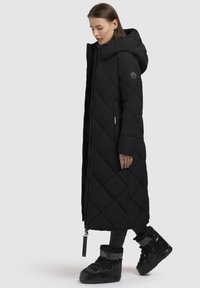 Sort lang quiltet pufferjakke med hætte, med et diagonalt mønster, sidelommer og en frontlynlås. Sammen med sorte støvler.