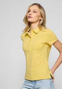 Żółta koszulka polo z krótkim rękawem, w slim fit, z kołnierzykiem i czterema guzikami na zapięciu. Posiada małe logo na piersi oraz boczne rozcięcia u dołu.