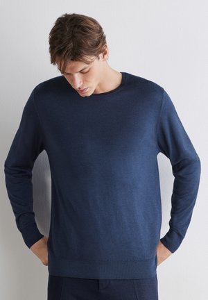 Ung mand iført en ensfarvet navyblå langærmet sweater, kigger ned med hænderne i lommerne, står foran en ensartet lys væg.