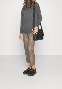 Frau in grauem Pullover, braunen karierter, cropped Hose, schwarzen Plateauslippern und mit schwarzer Schultertasche vor einfarbigem Hintergrund.