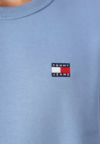 Felpa azzurra con collo a coste, dotata di un logo ricamato rettangolare in blu navy, bianco e rosso che reca la scritta "TOMMY JEANS."