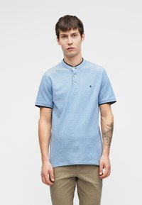 Vaaleansininen lyhythihainen polo-paita, jonka kauluksessa on napit, mustat reunat hihoissa ja pieni logo rinnassa. Sileä pinta, klassinen istuvuus.