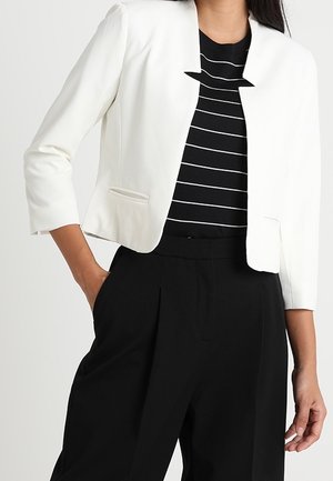 Mujer con blazer blanco corto sobre una blusa a rayas blancas y negras, combinada con pantalones negros de cintura alta, con la mano en el bolsillo.
