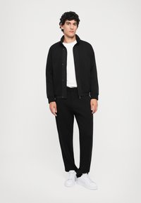Homme portant une veste zippée noire, un pantalon noir, un t-shirt blanc et des baskets blanches, se tenant devant un fond clair uni.