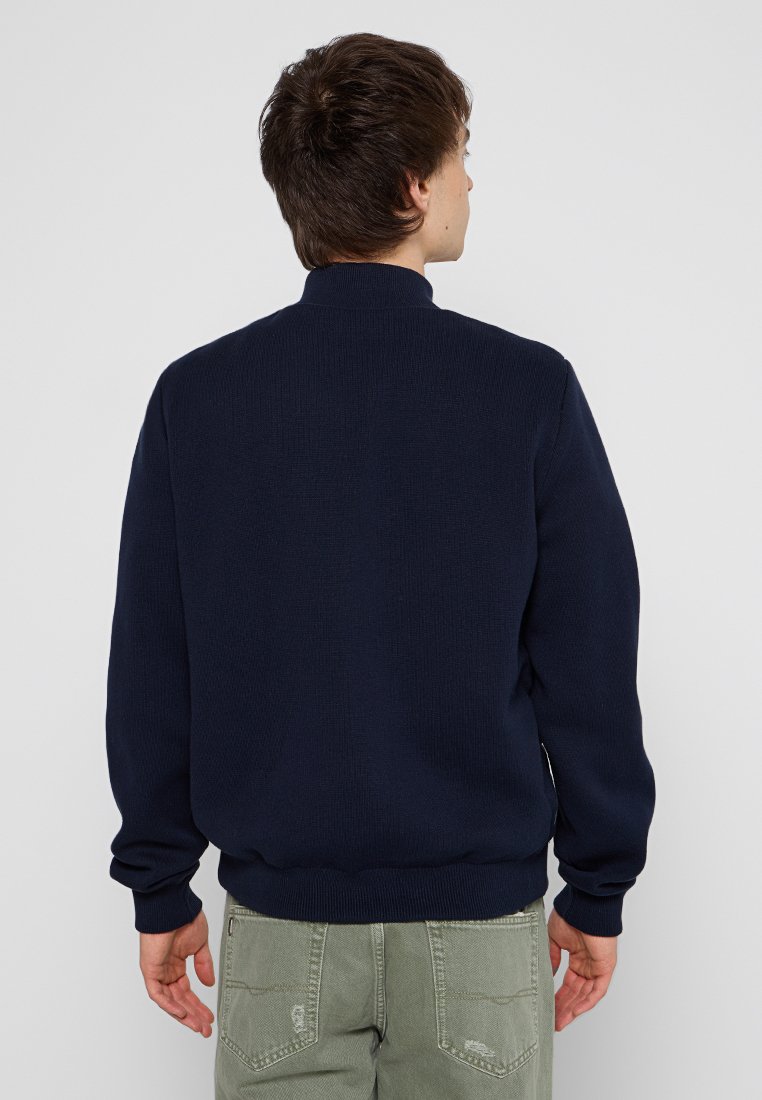 Dunkelblauer gestrickter Pullover mit hohem Kragen, gerippten Bündchen und Saum, getragen über hellgrünen Hosen. Schlichte Design ohne sichtbare Muster.