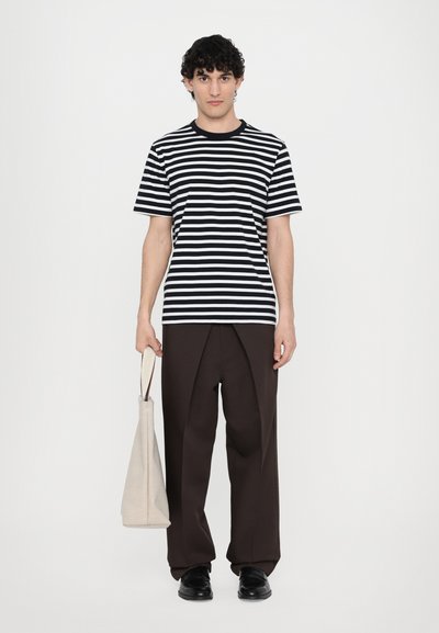 Camiseta de rayas en blanco y negro, de manga corta. Pantalones marrones holgados, bolsa beige de mano y zapatos negros completan el conjunto.