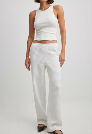 Kobieta w białej, bezrękawnikowej, prążkowanej krótkiej bluzce, wysokim stanem teksturowanych białych spodni o szerokich nogawkach oraz czarnych butach z ostrym noskiem.