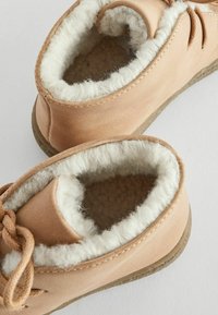 Chaussures slip-on en daim marron clair avec une douce doublure en fourrure crème et une semelle texturée beige. Dotées d'un laçage décoratif à l'avant.
