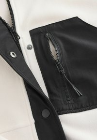 Fleecejacke mit einem cremefarbenen Körper, schwarzen Akzenten, einer Reißverschlusstasche und Druckknopfverschlüssen. Sie verfügt über eine glatte Textur und kontrastierende Materialien.