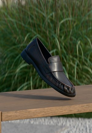 Musta nahkainen loafer-kengä, joka leijuu puupenkin yläpuolella, taustalla korkea vihreä ruoho on sumea.