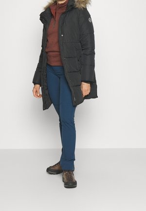 Persona con un abrigo acolchado negro con capucha y ribete de piel, suéter marrón, pantalones azules y zapatos de senderismo marrones, de pie frente a un fondo liso.