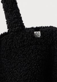 Zwarte faux fur totebag met een textuur, voorzien van een kort handvat en een klein merketiket met een logo op de bovenrand.