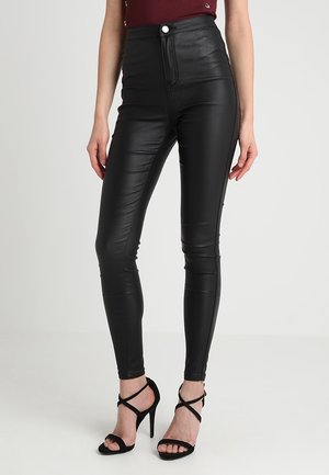 Femme portant un pantalon skinny taille haute en faux cuir noir et des sandales noires à talons hauts et fines lanières, debout sur un sol blanc.