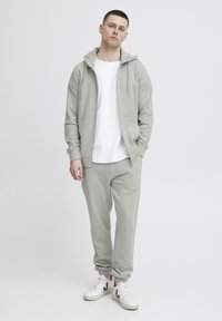 Felpa grigia chiara con zip frontale, tasche a marsupio e cappuccio con cordino. Abbinata a pantaloni della tuta coordinati e sneakers bianche.