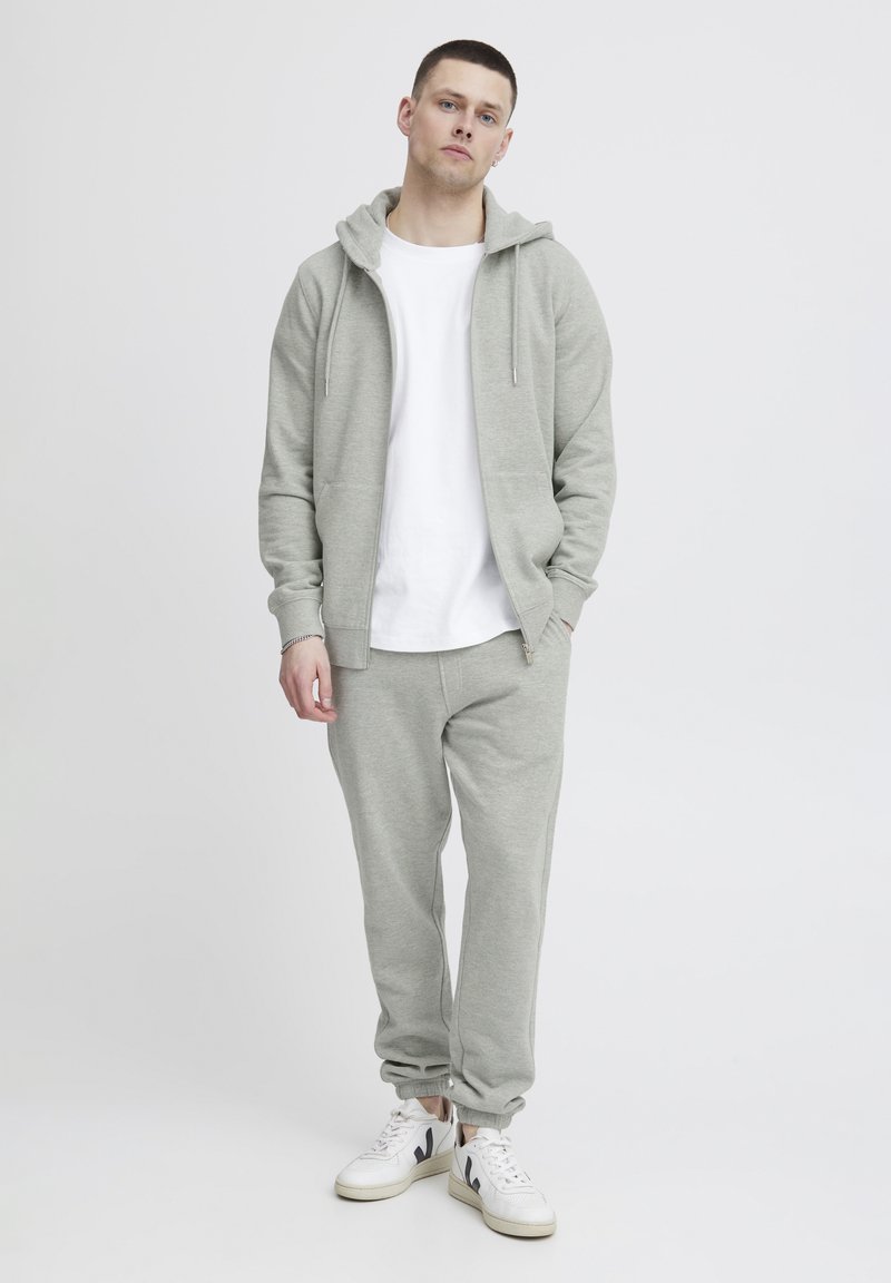 Felpa grigia chiara con zip frontale, tasche a marsupio e cappuccio con cordino. Abbinata a pantaloni della tuta coordinati e sneakers bianche.