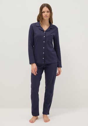 Femme portant un haut de pyjama boutonné à manches longues bleu marine avec col et un pantalon assorti, debout pieds nus devant un fond uni.