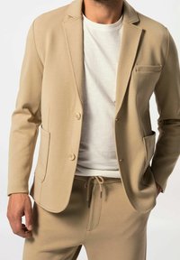 Beige sako vyrobené z elastické látky s dvěma předními kapsami a jedním knoflíkovým zapínáním, kombinované s bílým tričkem a odpovídajícími kalhotami.