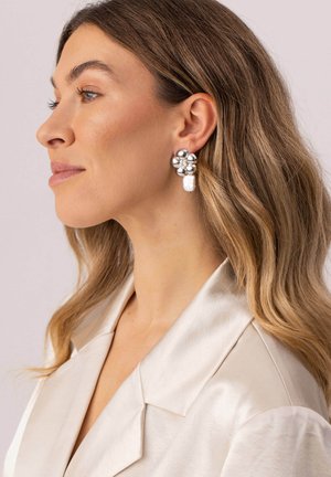 Frau mit welligem blondem Haar, die einen cremefarbenen Satinblazer und große silberne, blumenförmige Ohrringe mit Perltropfen trägt, nach links gewandt.