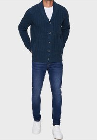 Donkerblauwe gebreide cardigan met een sjaalkraag, lange mouwen en een sluiting met vijf knopen. Draagt over een wit overhemd en gecombineerd met een blauwe spijkerbroek.