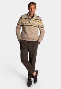 Beige gebreide sweater met een patroon op de borst featuring gele, bruine en grijze zigzags. Gepaard met donkere broek en marineblauwe sneakers.