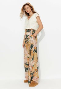 Haut blanc à col en V, pantalon floral ample avec des motifs pêche, vert et doré, doté d'une taille smockée et de poches, associé à des chaussures beiges.