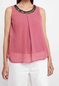 Ärmlös rosa topp med en pärlbesatt halsringning, plisserad framsida och en luftig, lätt silhuett. Kombinerad med vita byxor, visad framifrån.