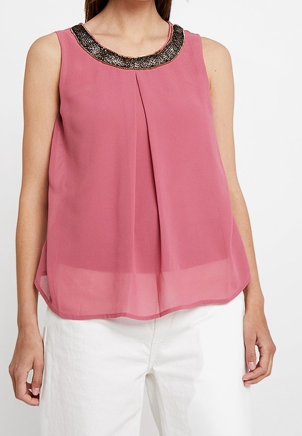 Ärmlös rosa topp med en pärlbesatt halsringning, plisserad framsida och en luftig, lätt silhuett. Kombinerad med vita byxor, visad framifrån.