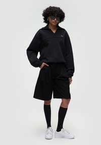Felpa nera a maniche lunghe e con collo alto, abbinata a pantaloni corti neri su misura. Il modello indossa calze nere fino al ginocchio e sneaker bianche.