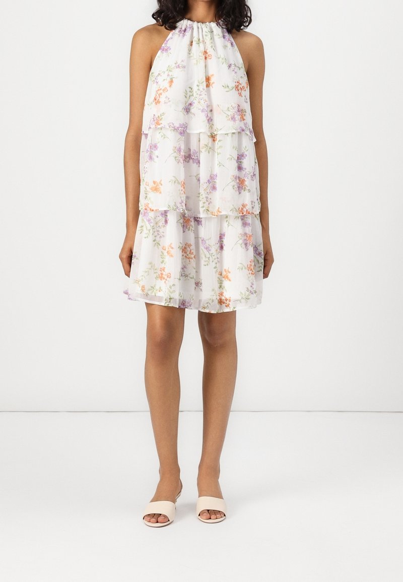 Femme portant une robe blanche fleurie sans manches à col licou avec jupe à volants et chaussures beige à bout ouvert, debout devant un fond blanc uni.