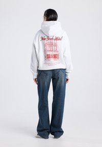 Persona con cabello corto y oscuro que lleva puesto un sudadera blanca con un gráfico rojo de "New York Hotel" y jeans azules, de pie contra un fondo liso.