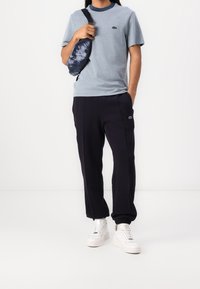 Person som bär en ljusblå t-shirt, svarta joggerbyxor, vita sneakers och håller en svart och blå batik-mönstrad axelväska framför en vit bakgrund.