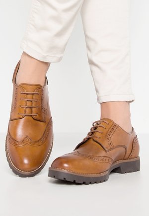 Brązowe skórzane buty eleganckie z wzorem brogu, z sznurowanym zapięciem i solidną gumową podeszwą. Noszone z podwiniętymi kremowymi spodniami.