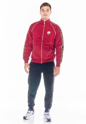 Un homme porte une veste de survêtement rouge zippée, ornée de bandes à motifs sur les manches, un pantalon de jogging foncé et des baskets blanches sur fond blanc.