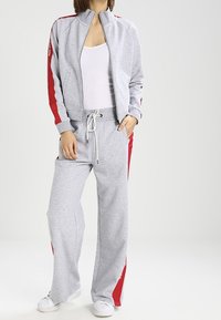 Grå idrottsdräkt med röda sidostreck. Dragkedjefodrad jacka, långa ärmar och byxor med dragsko. Vita sneakers fulländar utseendet.