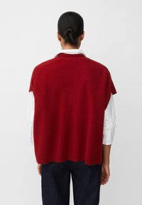 Gilet di maglia rossa con orlo a coste, design oversize, indossato sopra una camicia bianca a maniche lunghe. Texture liscia e silhouette leggermente drappeggiata.