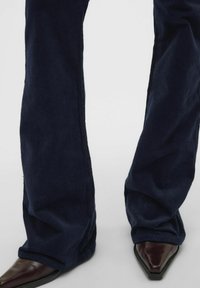 Pantalons en velours côtelé bleu marine évasés avec une texture en relief, présentant un ourlet légèrement effiloché. Associés à des chaussures à bouts pointus marron foncé.