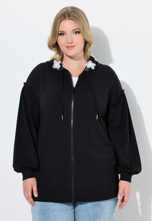 Lyshåret kvinde iført sort hættetrøje med lynlås og hvide snore med blomster samt lyseblå jeans, står foran en hvid baggrund.