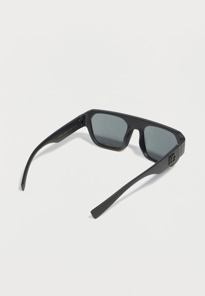 Μαύρα ορθογώνια γυαλιά ηλίου με σκούρα φακούς, χοντρά πλαϊνά και μικρή λεπτομέρεια λογότυπου στο πλάι, εμφανίζονται σε λευκό φόντο.