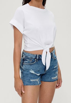 Kvinde iført hvid kortærmet crop top bundet ved taljen med opkrogede ærmer og slidte blå denimshorts.