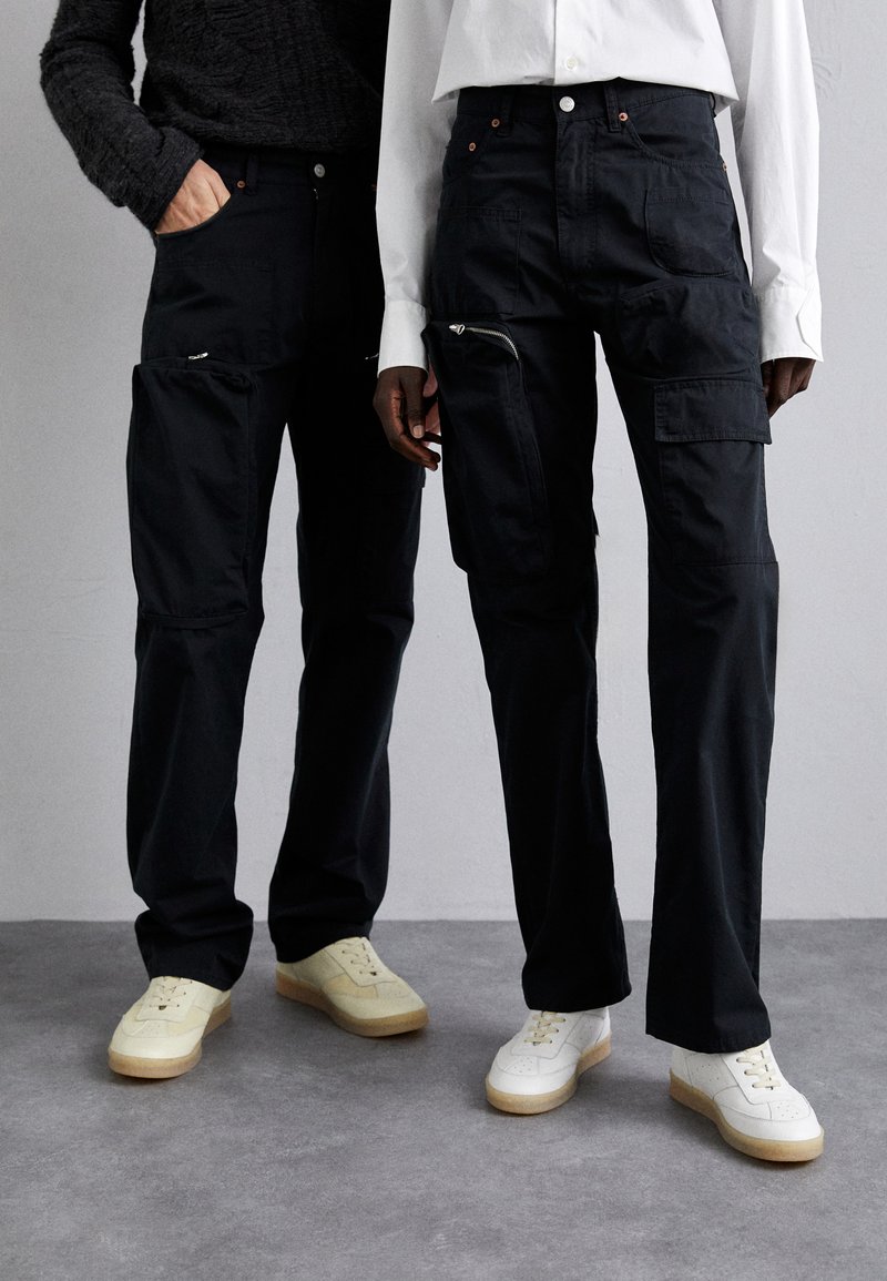 Black cargo pants with multiple pockets and zippers, paired with white sneakers. Fabric appears sturdy with a smooth texture.